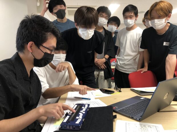 点字デバイスを操作する学生を囲んで複数の学生が見学している様子を映した写真